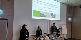 CHK 2023 Teresa Sartor, Dr. Jana York und Daniel Krüger (v.l.n.r.) bei ihrem Vortrag über Einheitliche Ansprechstellen für Arbeitgeber*innen. Sie stehen hinter einem grauen Metalltisch mit dem Rücken zu einer weißen Wand, auf der die Projektion der ersten Folie ihres Vortrags mit dessen Titel zu sehen ist.